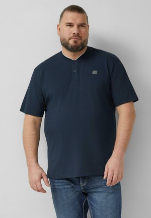 Mężczyzna z brodą, ubrany w granatową koszulkę Henley z krótkim rękawem i niebieskie dżinsy, stojący na jednolitym szarym tle.