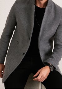 Cappotto grigio in misto lana con colletto alto, due tasche frontali e bottoni neri, abbinato a una camicia nera e un orologio da polso. Design slim fit.