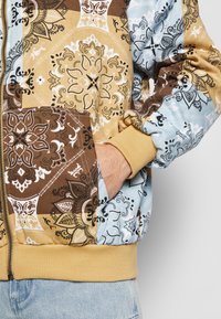 Jakna bomber s patchwork uzorkom koja sadrži cvjetne uzorke u smeđoj, bež i svijetloplavoj boji, rebraste manšete i zatvaranje na zip. Uključuje bočne džepove.