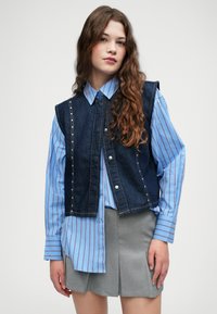 Džínová vesta s kovovými patentkami, nosená cez svetlomodrú pruhovanú košeľu. Sivá plisovaná sukňa dopĺňa outfit. Hladké textúry látok.