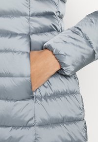 Mano dentro del bolsillo con cremallera de una chaqueta acolchada brillante de color gris claro con costuras horizontales.