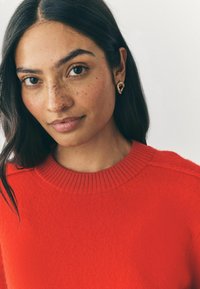 Maglione rosso a maglia con colletto a coste. Il modello ha capelli scuri e indossa orecchini d'oro a forma di orecchio. Pelle liscia con lentiggini.