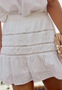 Jupe blanche en tissu texturé, présentant des plis verticaux et des accents en dentelle, avec un ourlet en volants pour plus de détails.