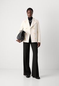 Dubbelzijdige crèmekleurige blazer met gouden knopen, gecombineerd met een zwarte coltrui, zwarte broek en een zwarte tas. Gladde textuur, op maat gemaakte pasvorm.