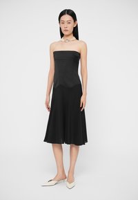 Čierne šaty bez ramienok s priliehavým živôtikom a rozšírenou sukňou. Hladká textúra, dĺžka do kolien, v kombinácii s bielymi topánkami s špičatou špičkou.