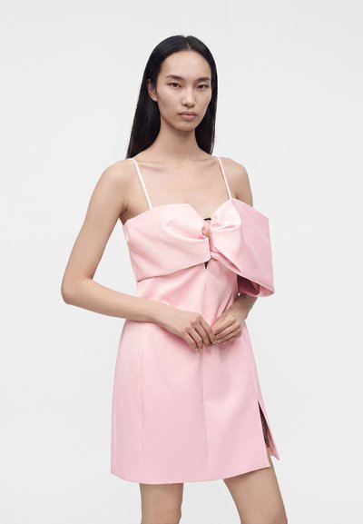 Femme aux longs cheveux noirs portant une robe rose sans manches avec un grand nœud sur la poitrine et une fente latérale.