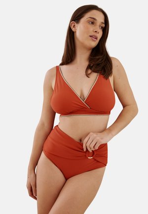 Femme portant un bikini couleur rouille avec un haut cache-cœur et un bas taille haute avec un anneau décoratif sur le côté, debout devant un fond uni.