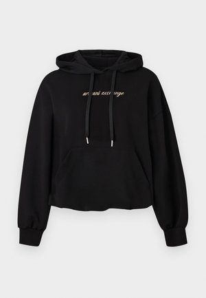 Femme debout, les mains dans les poches, portant un sweat à capuche noir avec texte blanc, un jogging noir assorti et des baskets blanches sur un fond uni.