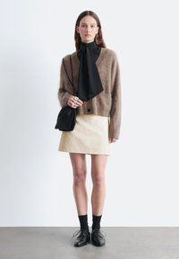 Brauner flauschiger Cardigan über einem schwarzen Oberteil mit einem Schal, kombiniert mit einem hellbeigen Rock und schwarzen Schuhen, mit glatten Texturen und subtilen Kontrasten.