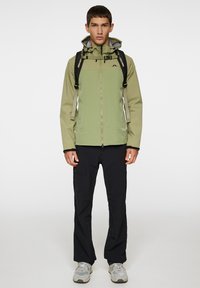 Lyst grøn vandtæt jakke med hætte, frontlynlås og sorte detaljer, båret over sorte bukser og grå sneakers, med en rygsæk.