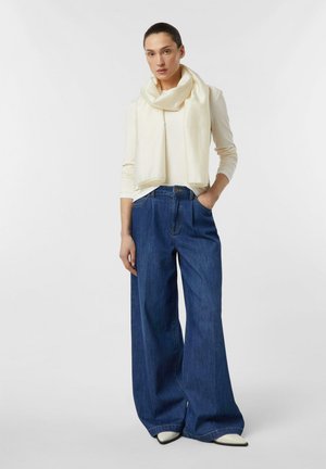 Woman wearing a long-sleeve cream top, matching scarf, wide-leg blue jeans, and white pointed-toe shoes, standing with one hand in pocket.