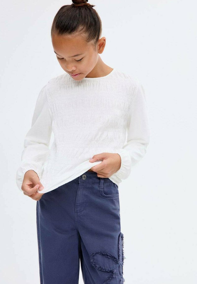 Maglietta bianca a maniche lunghe con parte anteriore testurizzata e scollo rotondo, abbinata a pantaloni blu navy con dettagli strappati lungo una gamba.