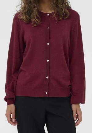 Femme portant un cardigan bordeaux à manches longues avec de petits boutons blancs et un pantalon noir, debout devant un fond uni.