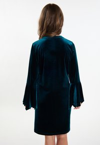 Tengerés színű bársonydressz harangujjakkal és kerek nyakkivágással, sima textúrával és térd fölé érő szabással, hátulról bemutatva.