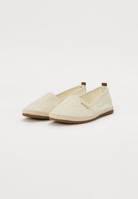 Ivory slip-on kengät, joissa on pitsinen päällyste, pyöreä varvas, teksturoitu kangas, beigestä ompelua ja ruskea yksityiskohta kantapäässä. Litteä kumipohja.