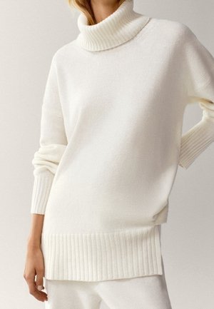 Femme portant un col roulé blanc côtelé à manches longues et un pantalon blanc assorti sur un fond uni.