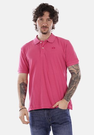 Uomo con capelli ricci che indossa una polo rosa acceso e jeans blu, mostra tatuaggi dettagliati su entrambi gli avambracci, in piedi con una mano in tasca.