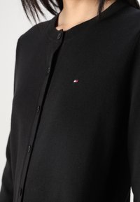 Zwarte cardigan met een ronde halslijn, knoopsluiting en een klein geborduurd logo op de borst. Zachte textuur en aansluitend ontwerp.