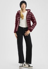 Burgunderfarbene Daunenjacke mit horizontalen Nähten, kombiniert mit schwarzen, hochgeschnittenen Jeans und schwarzen Sneakers. Model trägt eine schwarze Mütze.