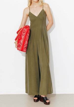 Femme en combinaison pantalon large sans manches de couleur vert olive, tenant un sac rouge à motifs, portant des sandales compensées noires.
