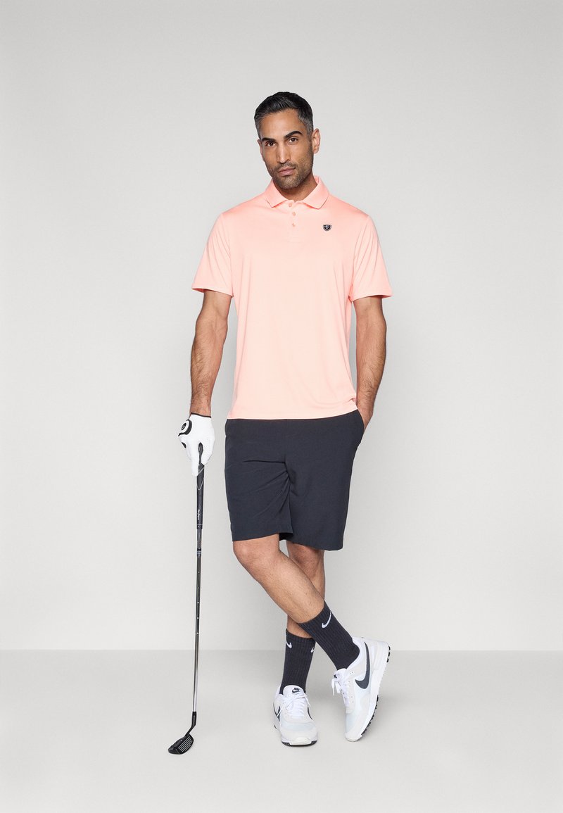 Polo rose clair à manches courtes, avec col et logo ; accompagné d'un short foncé, de baskets blanches et d'un club de golf.