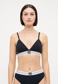 Black bralette with a deep V-neck, thin straps, and a wide white striped band featuring a logo at the front; smooth fabric texture.