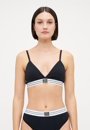 Black bralette with a deep V-neck, thin straps, and a wide white striped band featuring a logo at the front; smooth fabric texture.