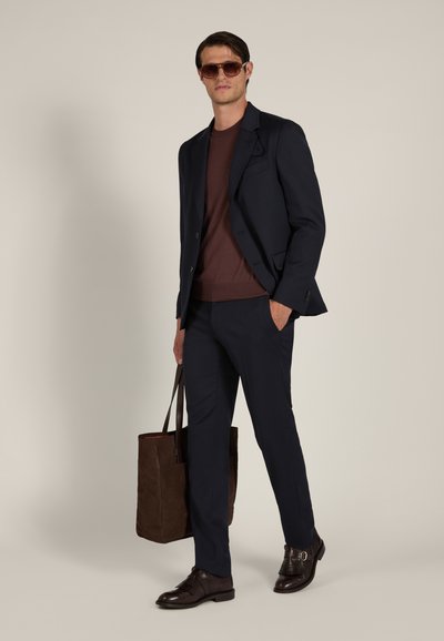 Man wearing dark suit, brown shirt, sunglasses, dark shoes, holding a large brown tote bag, standing against plain background.