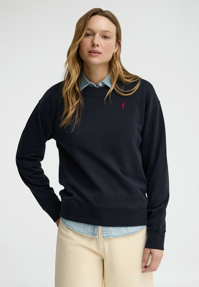 Vrouw met lang haar die een marineblauwe sweater draagt over een lichtblauw overhemd met knopen en beige broek, staand tegen een effen achtergrond.