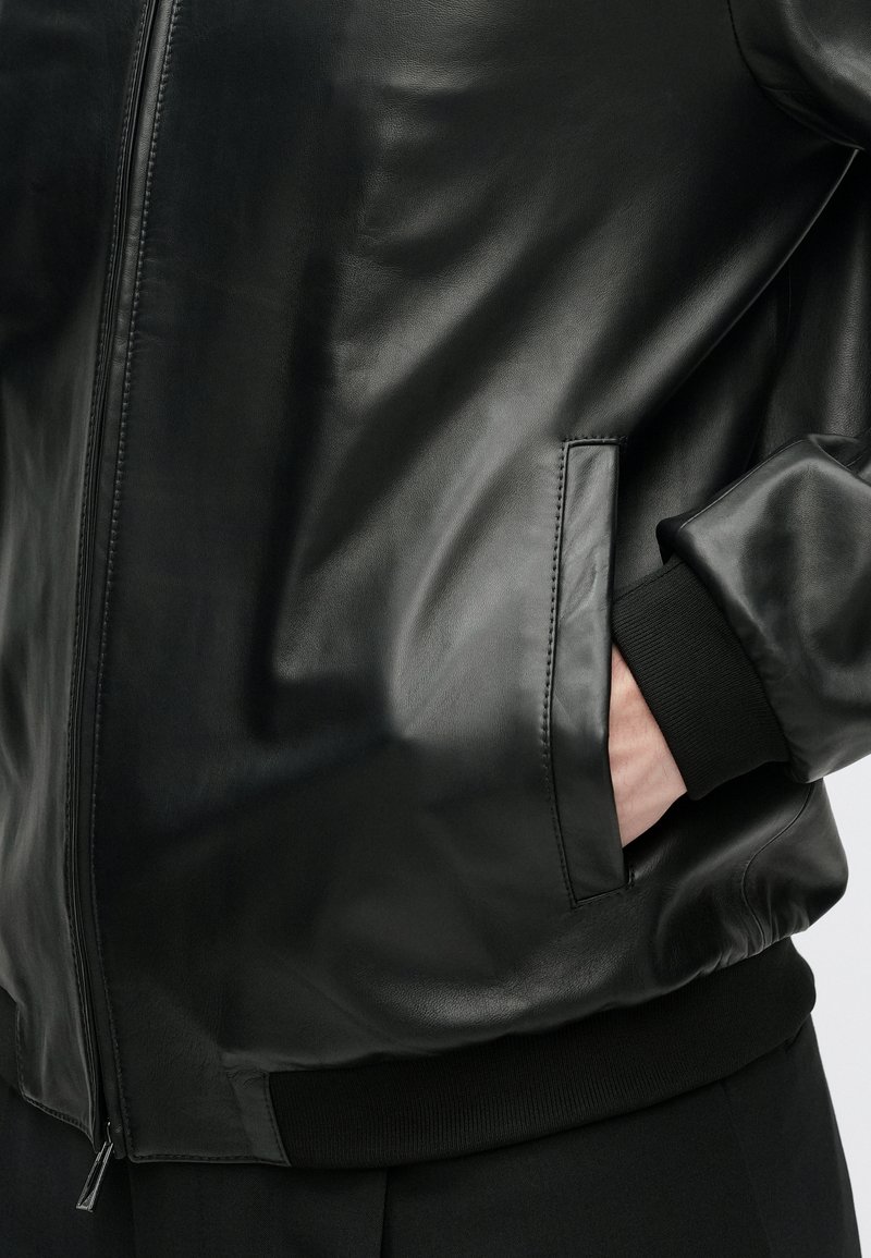 Veste aviateur en cuir noir avec une finition lisse, des poignets côtelés, des poches latérales et une fermeture éclair à l'ourlet.