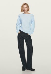 Donna con capelli biondi corti, che indossa un maglione azzurro chiaro, jeans larghi scuri e scarpe nere, in piedi con le mani in tasca su uno sfondo neutro.