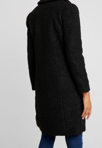 Manteau texturé noir de longueur moyenne, avec un grand col, des manches longues et une coupe droite, porté par-dessus un jean foncé.