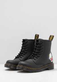 Černé šněrovací boty s žlutým šitím a postavou Hello Kitty na vnější straně paty, na jednobarevném pozadí.