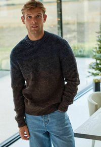 Pulover tricotat texturat întunecat, cu guler la baza gâtului, având un design gradient de la maro închis la maro deschis, asortat cu blugi din denim de un albastru deschis.