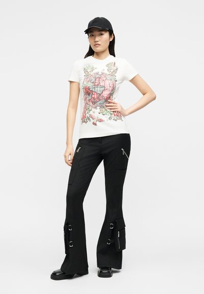 Γυναίκα με μαύρο καπέλο, λευκό μπλουζάκι με γραφική εκτύπωση γήινου σφαίρας, μαύρο παντελόνι cargo με αγκράφες και φερμουάρ, στέκεται με το χέρι στη μέση.