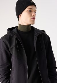 Must have kerge must jope kapuutsiga ja tekstureeritud paneeli disainiga. Kantud tumeda särgi üle, millel on soonikkoes roheline kõrge kaelus, ja must beanie.