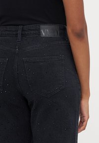 Jeans de ganga pretos com uma superfície texturizada apresentando pequenos strass. Dois bolsos traseiros e uma etiqueta preta de marca na cintura.