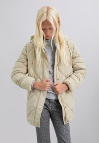 Blond persoon in een beige gewatteerde jacket over een grijze trui en patroon broek, die de rits van het jacket sluit, tegen een effen achtergrond.