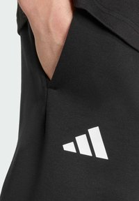 Sort stof med en glat tekstur, der har et hvidt Adidas-logo i et diagonalt mønster på siden. Lomme synlig ved sømmen.