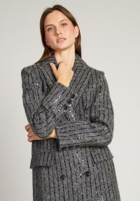 Abrigo de mezcla de lana gris, de doble botonadura, con rayas verticales de lentejuelas, cuello con muesca y bolsillos delanteros. Presenta botones negros y una textura de tela.