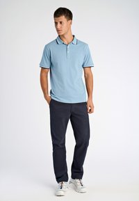 Jasnoniebieska koszulka polo z kołnierzykiem i dwoma guzikami, akcentowana granatowymi paskami. Noszona z ciemnymi spodniami i białymi sneakersami.