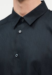 Κοντινό πλάνο ενός ατόμου που φοράει μαύρο πουκάμισο με κουμπιά, με διακριτό υφαντό μοτίβο και κλασικό γιακά.