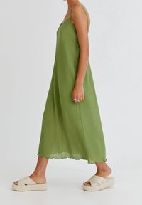 Kobieta spacerująca w bezrękawnej, zielonej, teksturowanej sukience midi z falbaniastym dołem i beżowych sandałach na platformie, ukazująca profil boczny od ramion w dół.