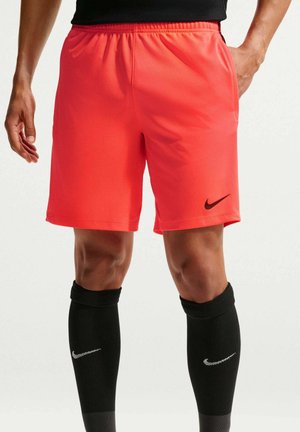 Mies, jolla kirkkaanpunaiset Nike-shortsit ja mustat Nike-polvisukat, seisoo yhdellä kädellä taskussa yksiväristä vaaleaa taustaa vasten.