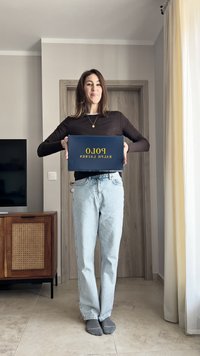 Sötét, hosszú ujjú felsőt és világoskék farmert viselő személy, aki egy sötétkék dobozt tart, amelyen sárga betűkkel szerepel a "POLO RALPH LAUREN" felirat.