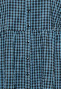 O tecido de padrão xadrez azul e preto apresenta um design em camadas com pregas verticais, terminado com botões pretos ao centro.