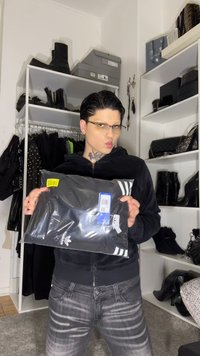 Čierna mikina Adidas v priehľadnom plastovom obale, s bielymi pruhmi na okrajoch, s viditeľnou modrou nálepkou a žltou cenovkou.