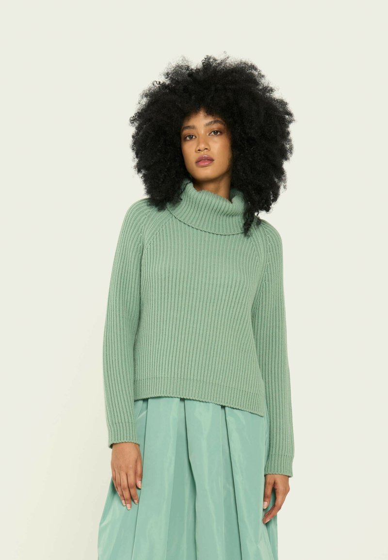 Mörkgrön stickad tröja med hög krage och ribbad textur, med långa ärmar och avslappnad passform. Kombinerad med en ljusblå kjol.