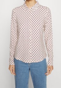 Camicia a maniche lunghe in tessuto beige chiaro con motivo floreale bordeaux, colletto con bottoni e orlo curvo, abbinata a jeans in denim blu.
