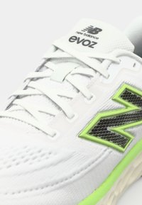 White New Balance sneaker with a textured upper, gray laces, and a vibrant green accent on the side. Features a soft, cushioned sole.